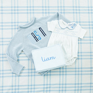 A charming baby gift set featuring a personalized blue sweater, a white onesie and a pouch.