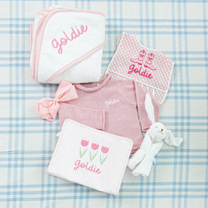 A personalized baby gift featuring a hooded towel, bib, striped onesie, pink bow.